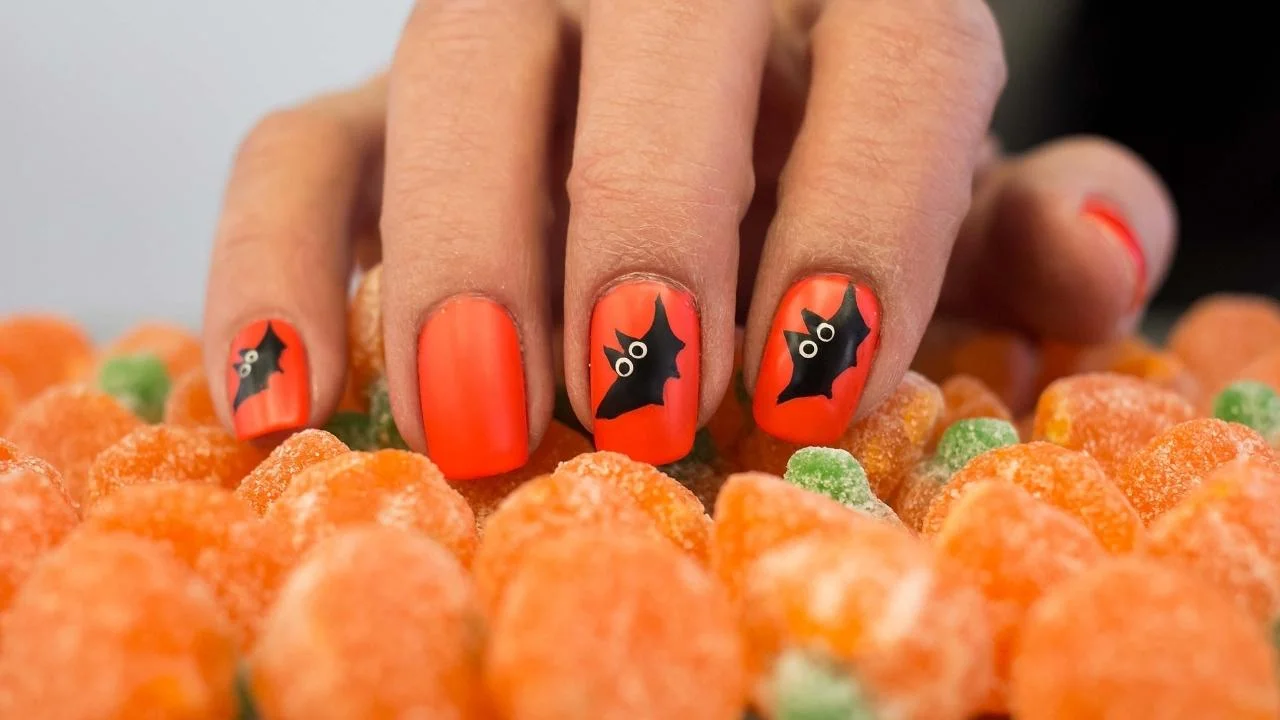 Paznokcie na Halloween pomalowane na pomarańczowo i z przyklejonymi ozdobami w kształcie nietoperzy
