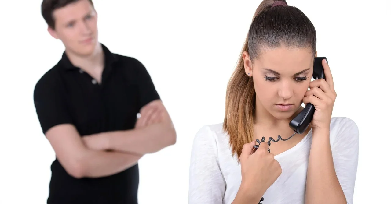 Kobieta rozmawiająca przez telefon, za nia mężczyzna z założonymi rękami